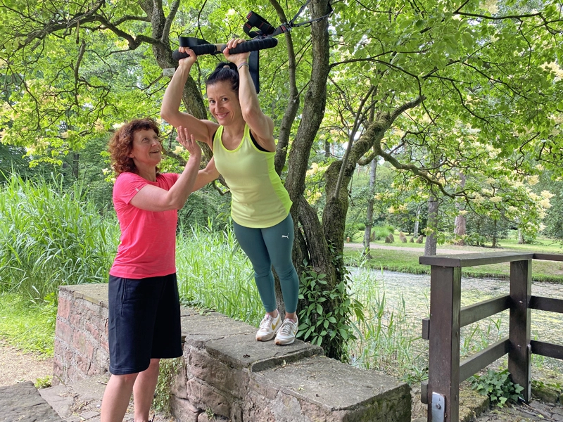 Kundin trainiert mit Suspension Trainer an einem Baum, unterstützt von Trainerin – funktionelles Outdoor-Training für Kraft, Koordination und Beweglichkeit