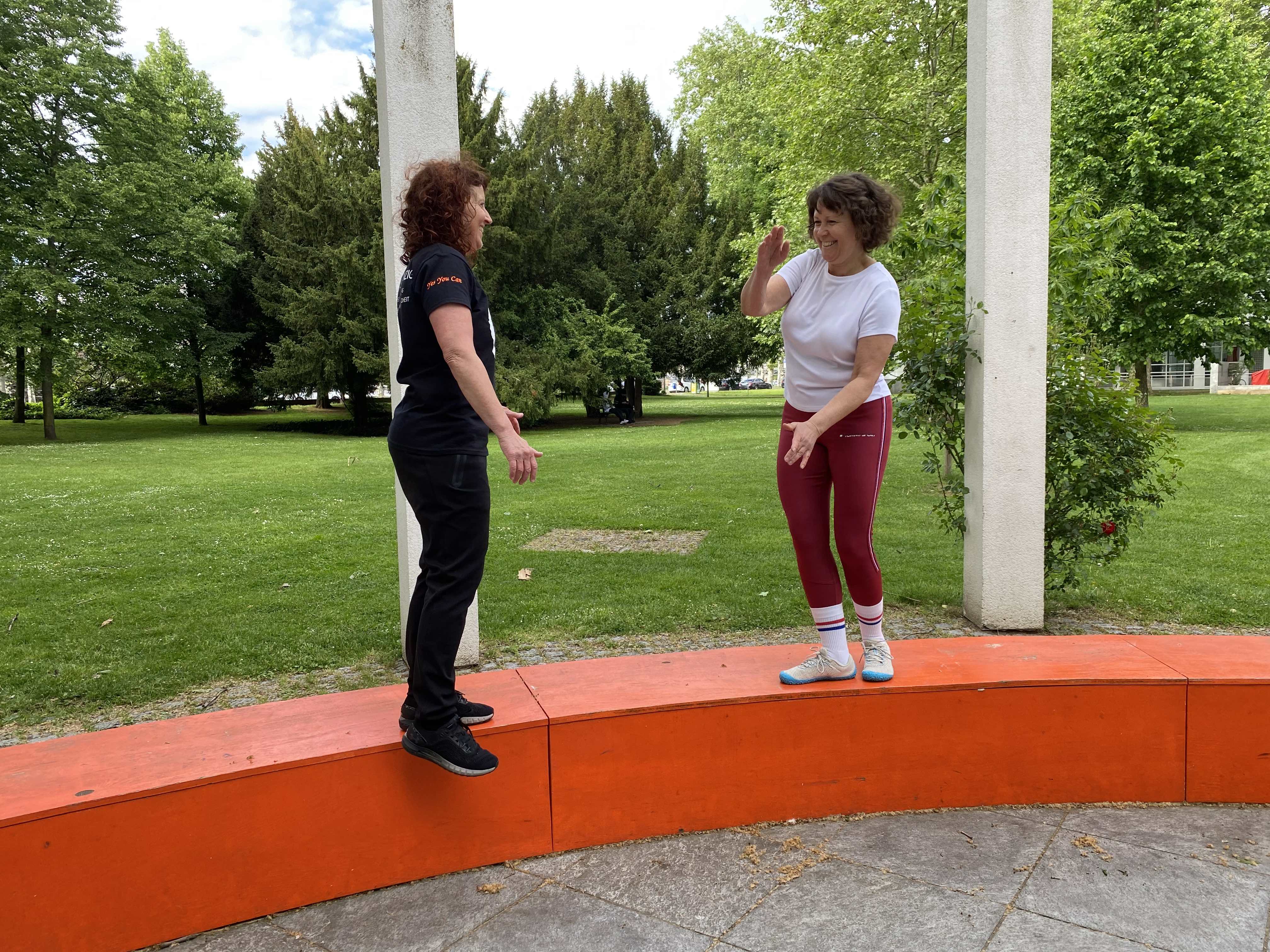 Trainerin und Kundin balancieren lachend auf einem Mäuerchen – Personal Training mit Spaß und Gleichgewicht.