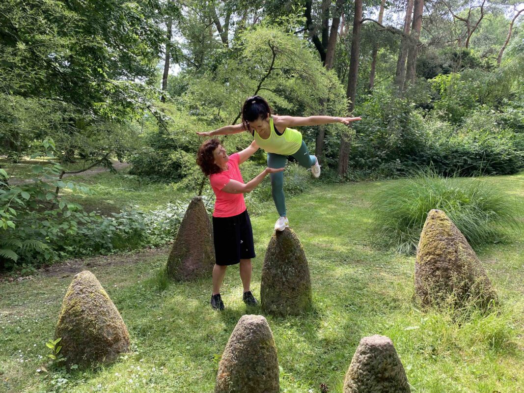 Personal Trainerin Marion Kinzig leitet Kundin bei einer Standwaage auf einem Stein im Grünen an – So geht Progression im funktionellen Training.