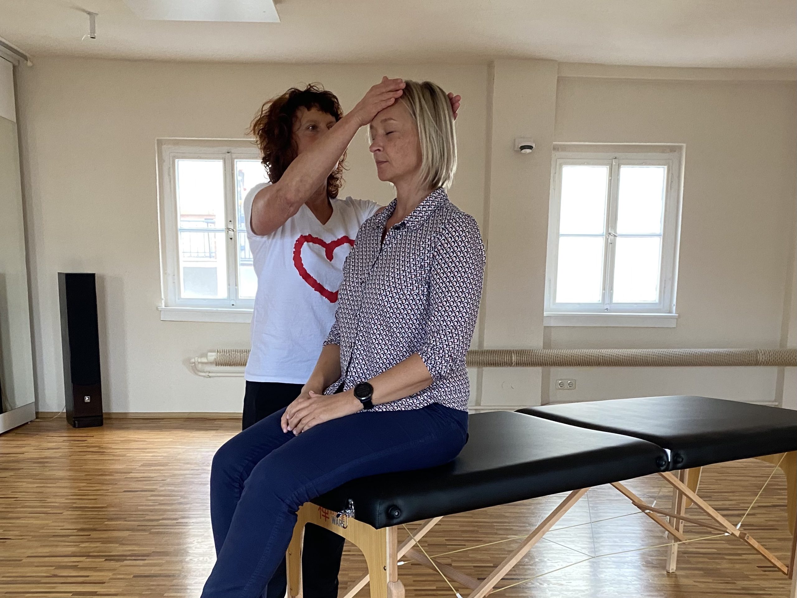 Zwei Personen bei einer Emotional‑Stress‑Release‑Anwendung; eine sitzt, die andere hält sanft Stirn und Hinterkopf.