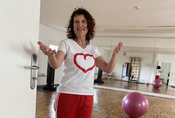 Marion steht lächelnd in der Tür ihres Frei‑Raums und macht eine einladende Geste. Im Hintergrund sind eine Spiegelwand und einTrainingsball zu sehen.